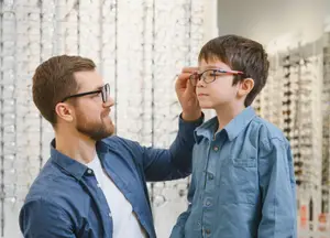 Male optician fitting glasses on young boy inside eyewear store.