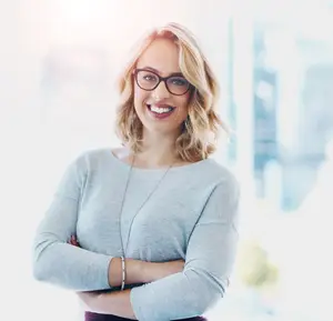 Smiling woman with blonde hair wearing glasses in modern office setting.